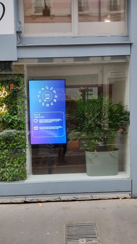 Écran vitrine affichant un horoscope numérique dans un commerce avec décor végétal.